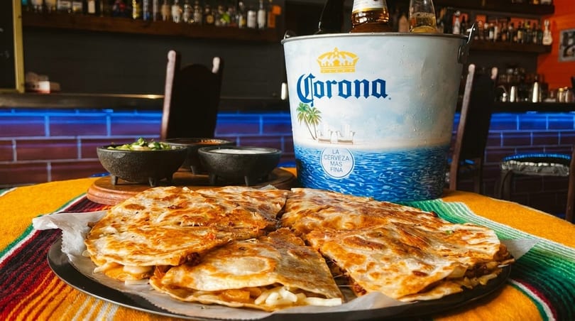 Quesadillas, dips, and a Corona beer bucket on a colorful striped blanket in a restaurant.