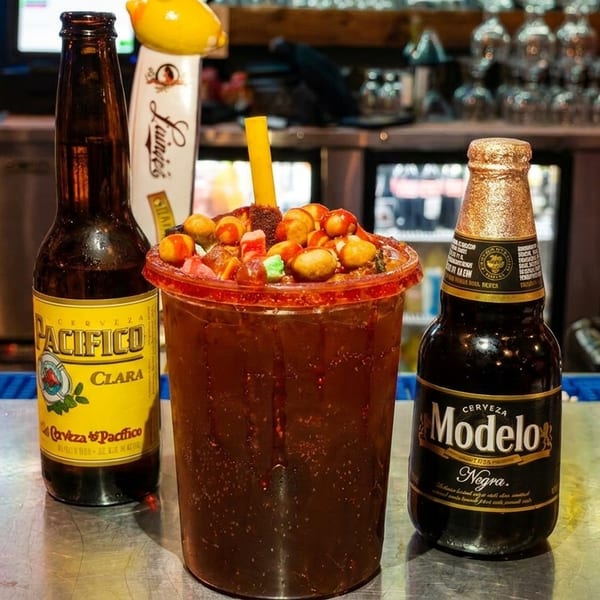 A Michelada drink with candy and peanuts, flanked by Pacifico Clara and Modelo Negra beer bottles.