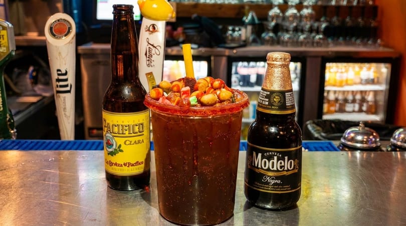 A Michelada drink with candy and peanuts, flanked by Pacifico Clara and Modelo Negra beer bottles.