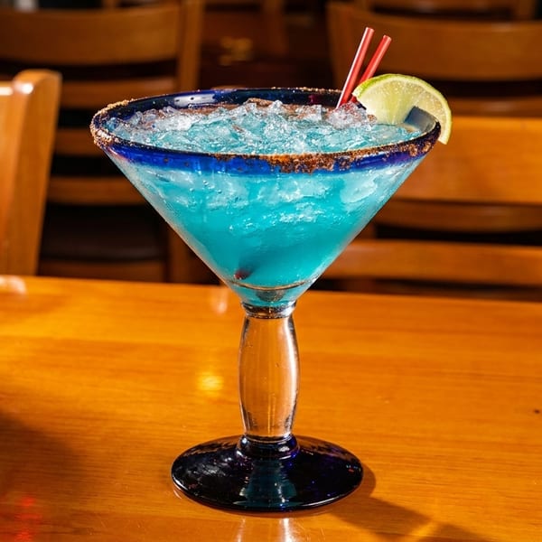 Blue margarita with ice, lime, and red straws on a wooden table with chairs in the background.