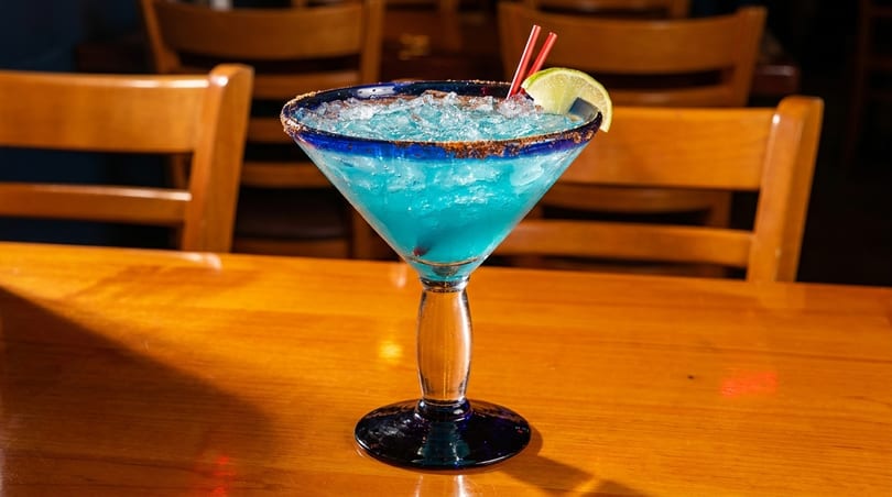 Blue margarita with ice, lime, and red straws on a wooden table with chairs in the background.