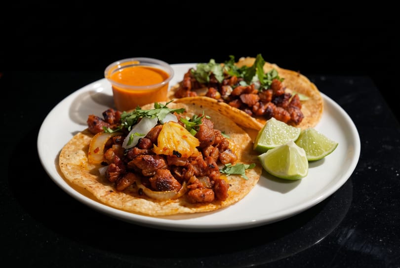 Two al pastor tacos with pineapple, onions, cilantro, lime wedges, and orange sauce on a white plate.