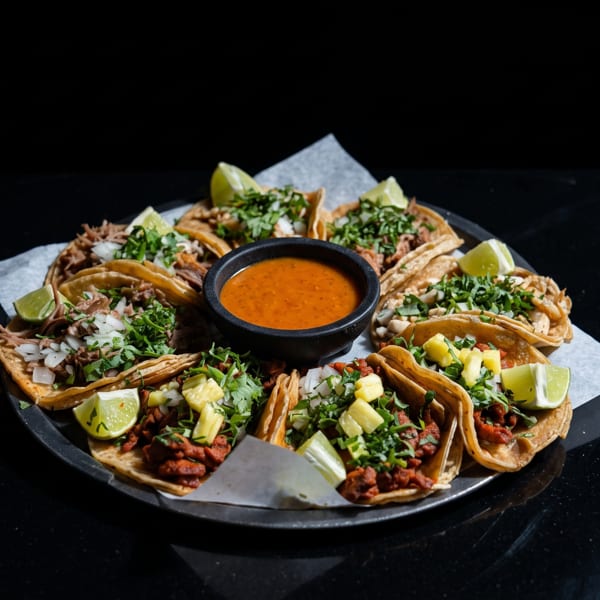 A platter of various tacos with lime wedges and a bowl of salsa on a dark surface.