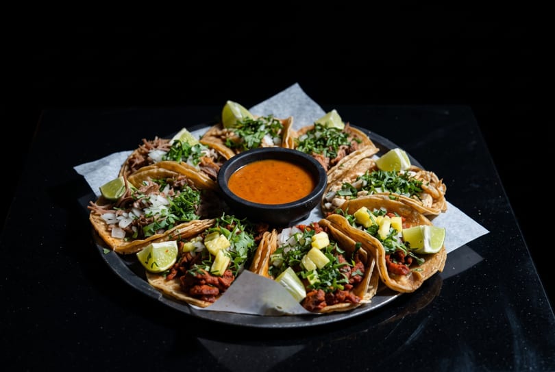 A platter of various tacos with lime wedges and a bowl of salsa on a dark surface.