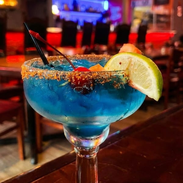Blue margarita with a chili-lime rim, lime, and cherries, on a dark wooden table in a restaurant.