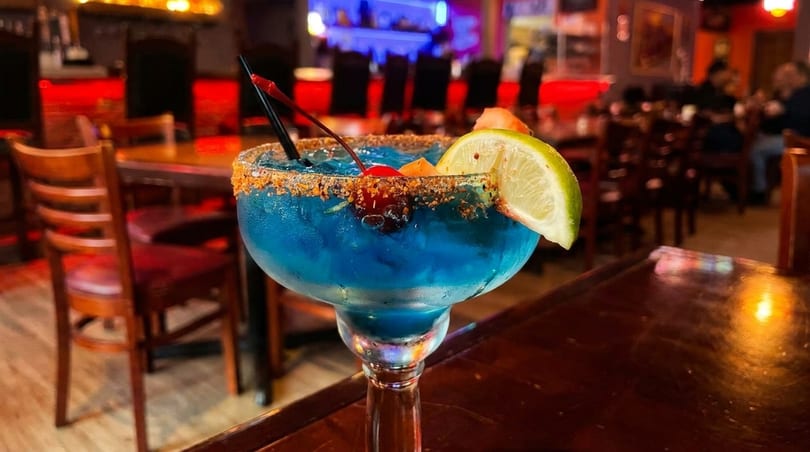 Blue margarita with a chili-lime rim, lime, and cherries, on a dark wooden table in a restaurant.