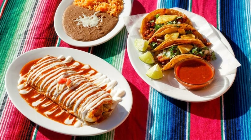 A burrito, tacos, rice, and beans on a colorful striped tablecloth.