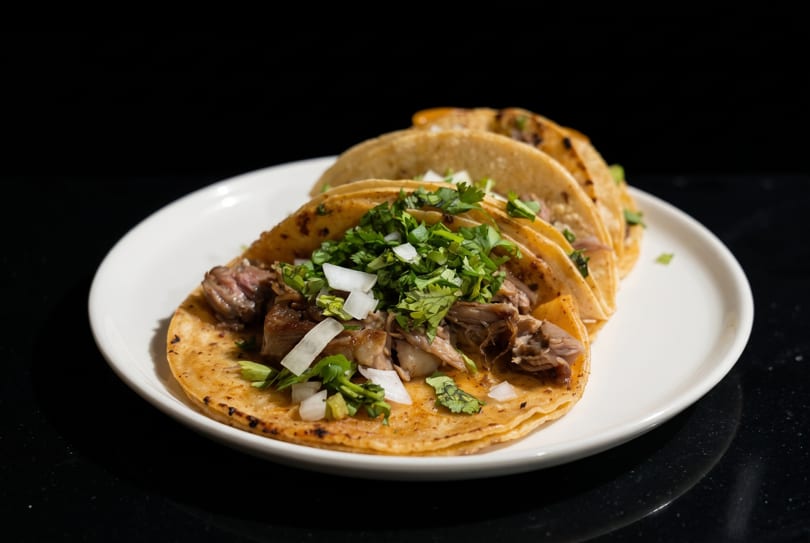 Three street tacos with meat, cilantro, and diced onions on a white plate.