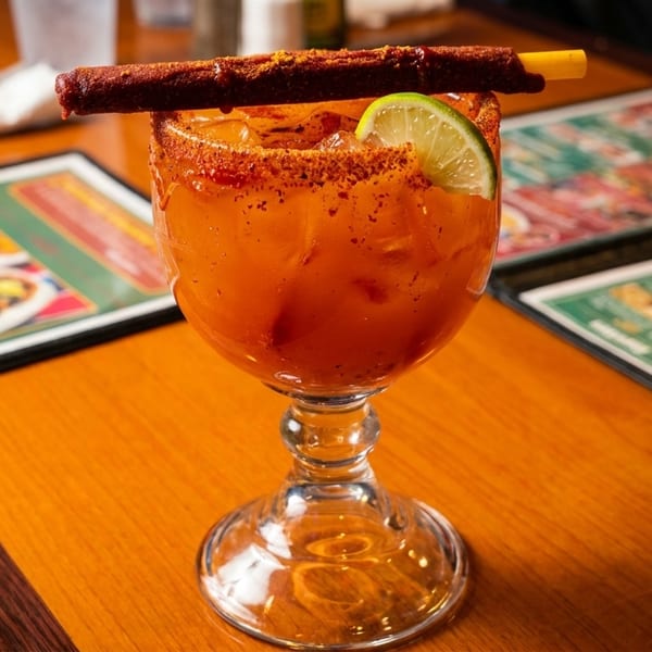 A vibrant orange cocktail with a lime wedge and a chili-dusted candy straw on a wooden table.