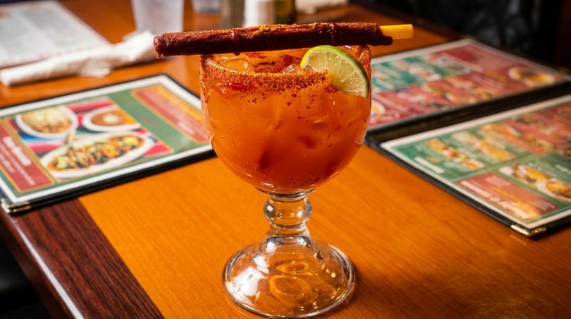 A vibrant orange cocktail with a lime wedge and a chili-dusted candy straw on a wooden table.