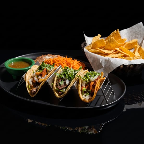 Three tacos in metal holders, with sides of refried beans, shredded carrots, salsa, and tortilla chips.