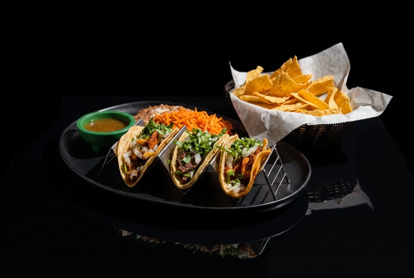 Three tacos in metal holders, with sides of refried beans, shredded carrots, salsa, and tortilla chips.